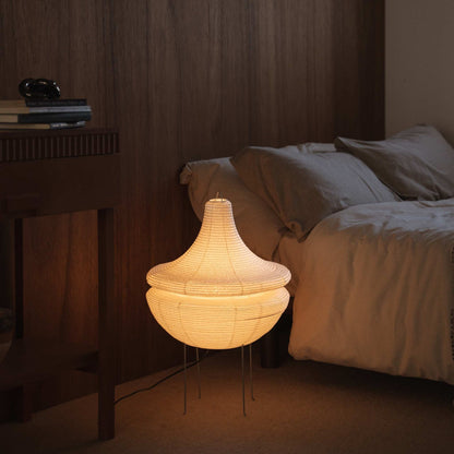 "Lampe sur pied japonaise illuminée Gemini 02 en papier, diffusant une lumière douce et chaude, posée au sol à côté d'un lit dans une chambre japonaise"