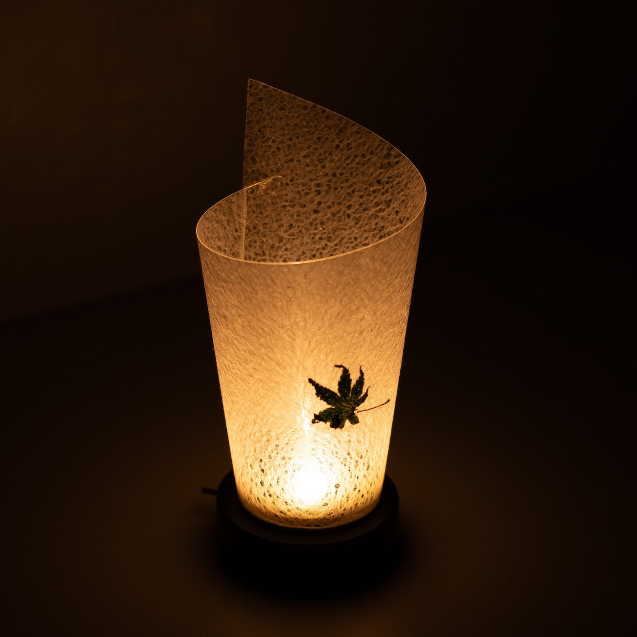 "Lampe de table japonaise en papier « Maki » feuille Momiji, blanc, papier Mino Washi - vue en plongée d'une lampe Japandi allumée montrant son design roulé."