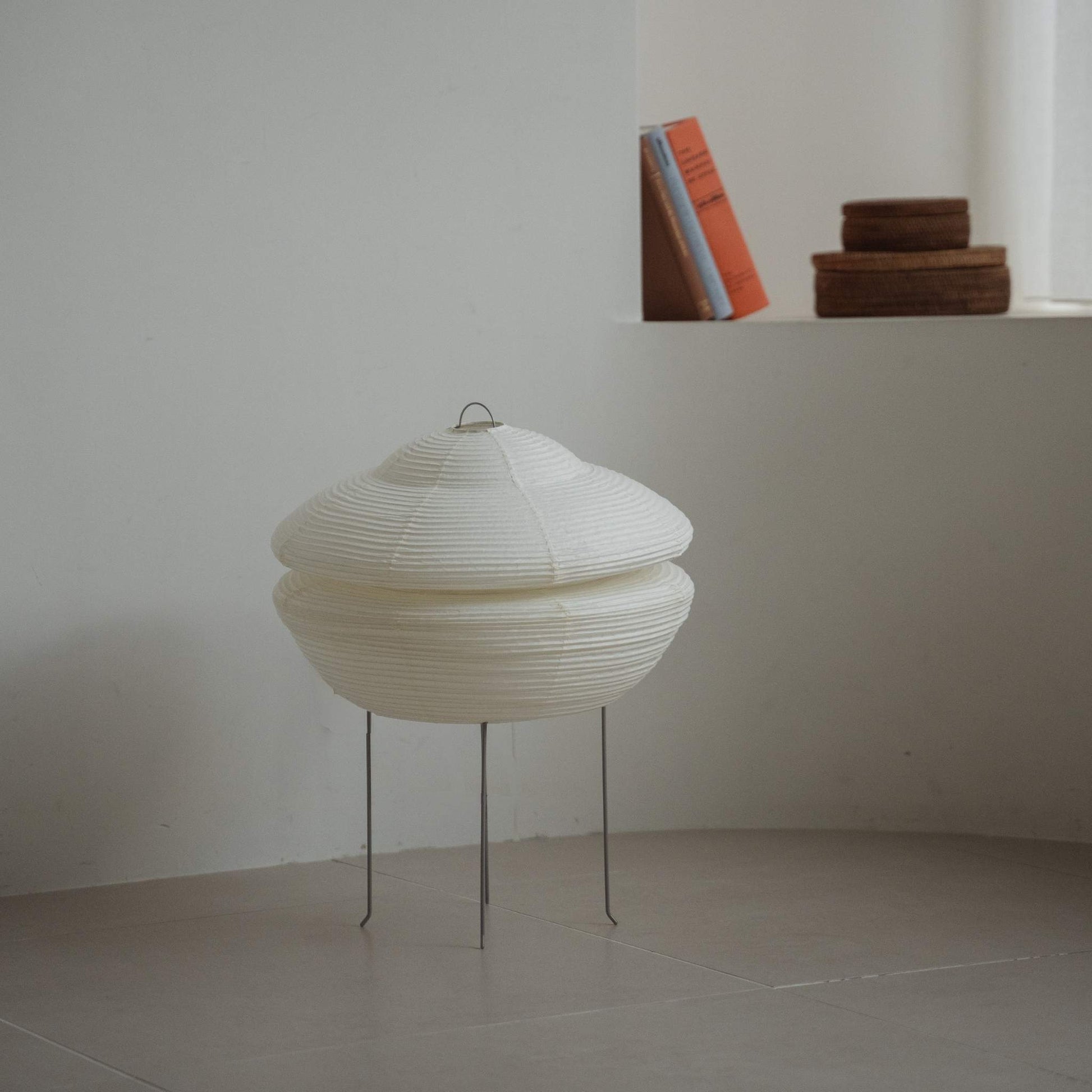 "Lampe japonaise moderne en papier Gemini 01 posée sur un sol carrelé dans une pièce blanche de style Japandi avec des livres sur une étagère derrière"
