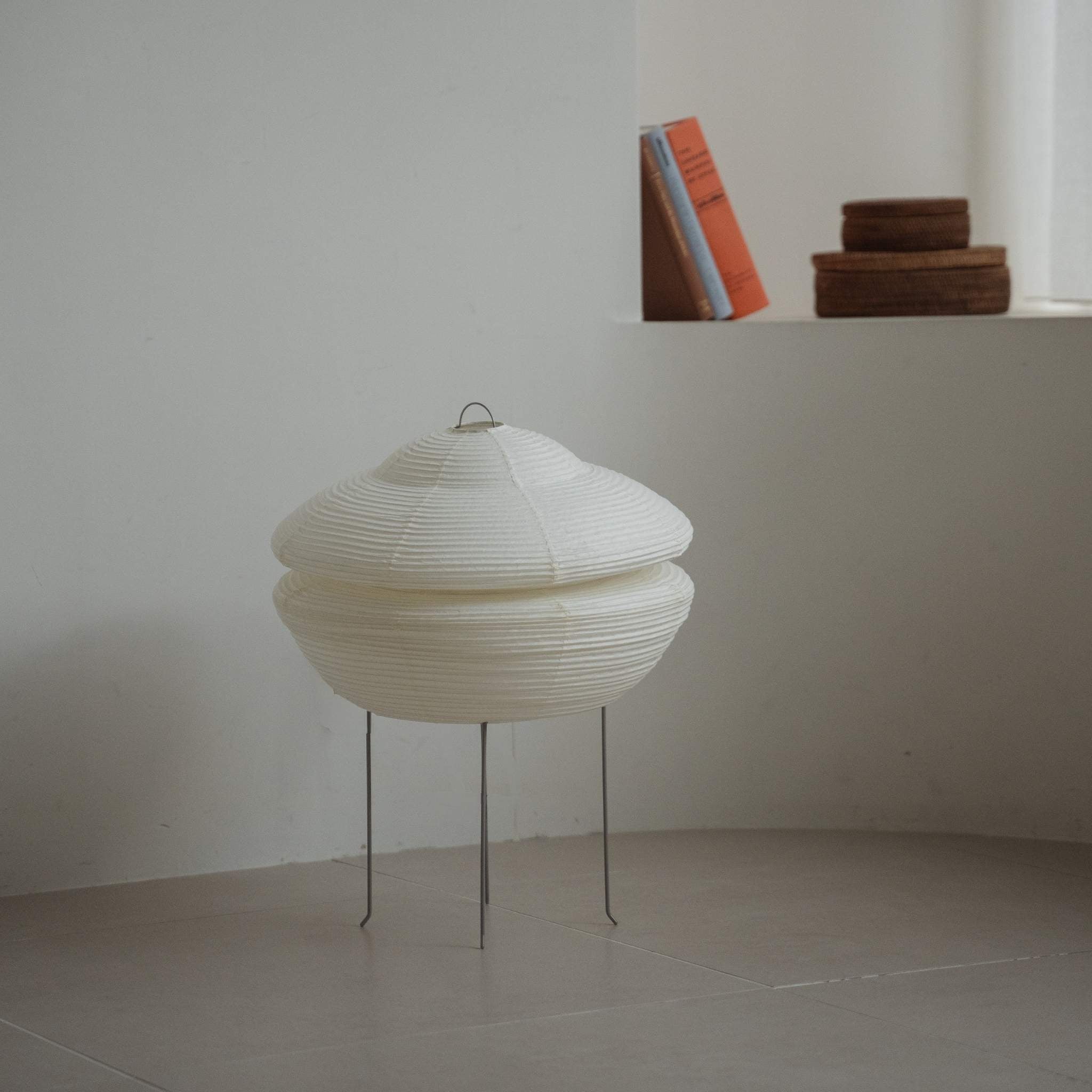 "Lampe japonaise moderne en papier Gemini 01 posée sur un sol carrelé dans une pièce blanche de style Japandi avec des livres sur une étagère derrière"