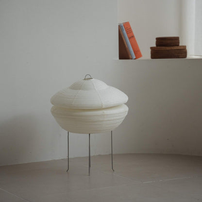 "Lampe japonaise moderne en papier Gemini 01 posée sur un sol carrelé dans une pièce blanche de style Japandi avec des livres sur une étagère derrière"