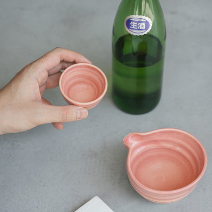 A hand holds a pink Japanese sake cup over a grey surface, with the matching sake pitcher (katakuchi) nearby.