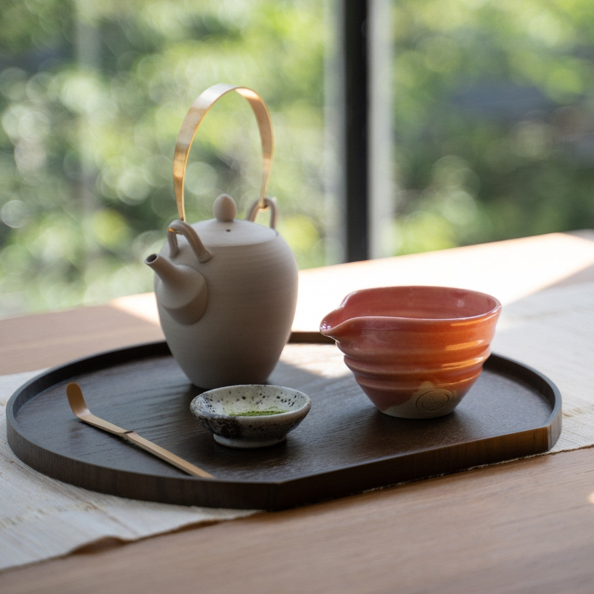 Pink Japanese matcha bowl with spout and tea tools on a dark tray by a sunny window, set for a peaceful authentic tea time.