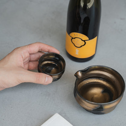 Eine Hand hebt vorsichtig einen handgefertigten Sakebecher (Ochoko) von einer Betonoberfläche, um einen Schluck zu nehmen, mit der Sake-Karaffe und Flasche in der Nähe.