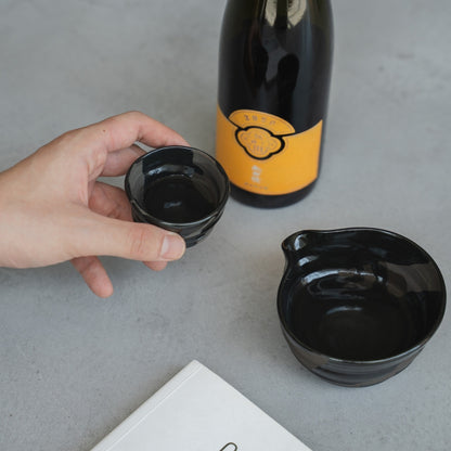 hånd holder et håndkastet Sumi Black japansk sakeglas (ochoko), på et gråt bord med en sakeflaske, til mindful sake-smagning.