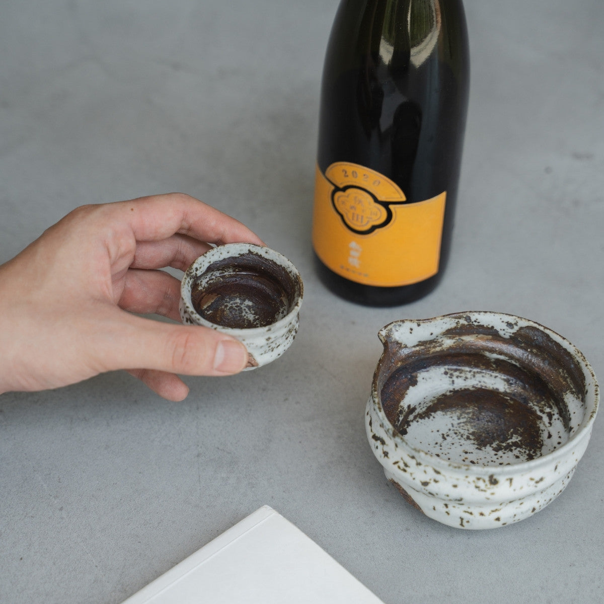 A hand holding an uneven white Zui Japanese sake cup (ochoko) by a table, for savoring premium sake in the evening
