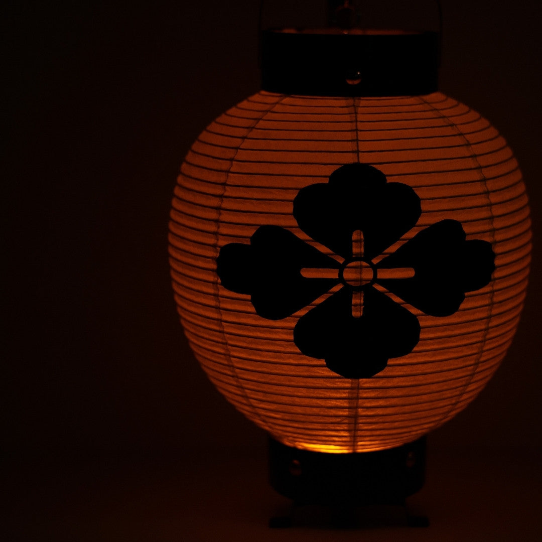 Close-up van een Japanse washi-papieren lantaarn met een bloemenwapenschild dat oplicht in een donkere ruimte voor een rustige sfeer.
