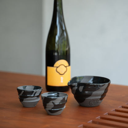 Sumi sort keramik japansk sake-sæt, Katakuchi karaffel & 2 kopper, på et træbord, nær en flaske sake, til mindful aftensritualer.