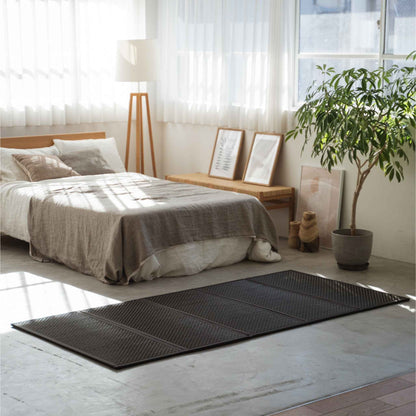 Enkel sort tatami-måtte placeret på et betongulv i et solbeskinnet rum med en minimalistisk japansk indretningsstil.