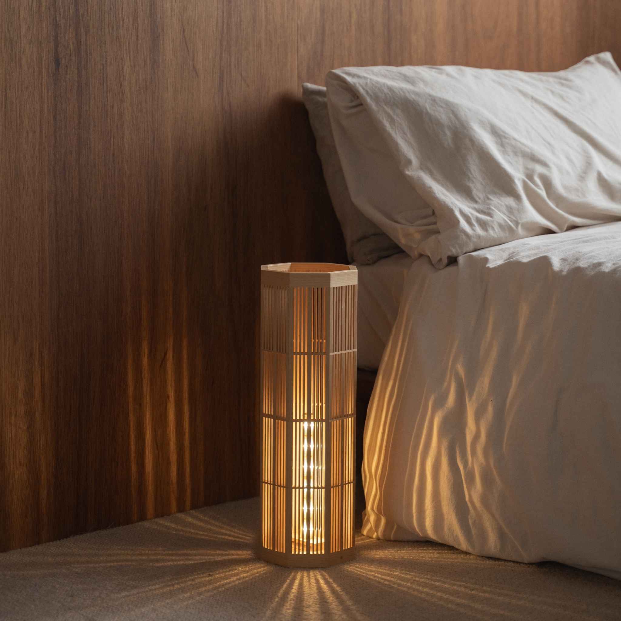 "Lampadaire japonais en bois « SAN » Grand, placé à côté d'un lit, diffusant une lumière d'ambiance chaleureuse pour une nuit paisible."