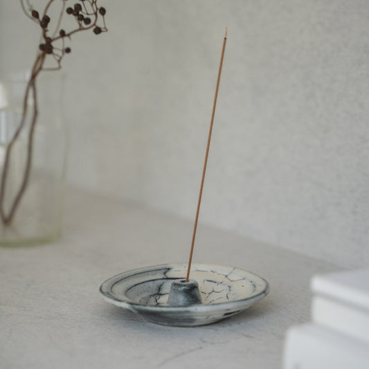 Witte en grijze Japanse wierookhouder met een brandende stok op een lichte tafel, wat sereniteit creëert voor ochtendmeditatie.