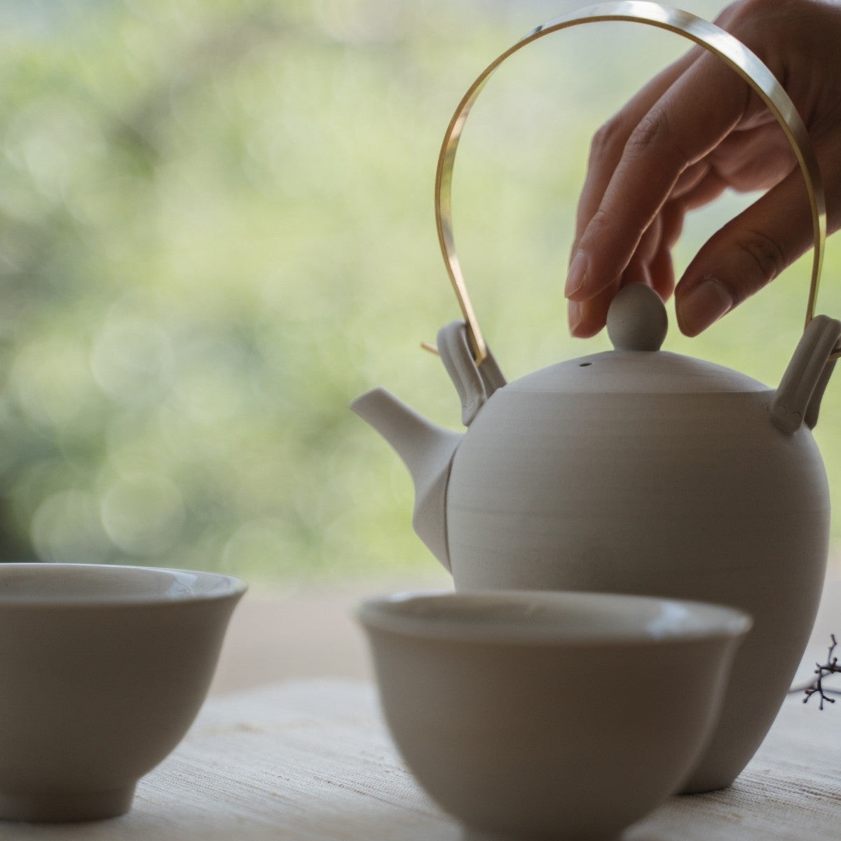 Infusion de thé à la main avec une théière japonaise blanche de type kyusu, près d'une fenêtre lumineuse, pour les amateurs de thé débutant leur matinée en pleine conscience.