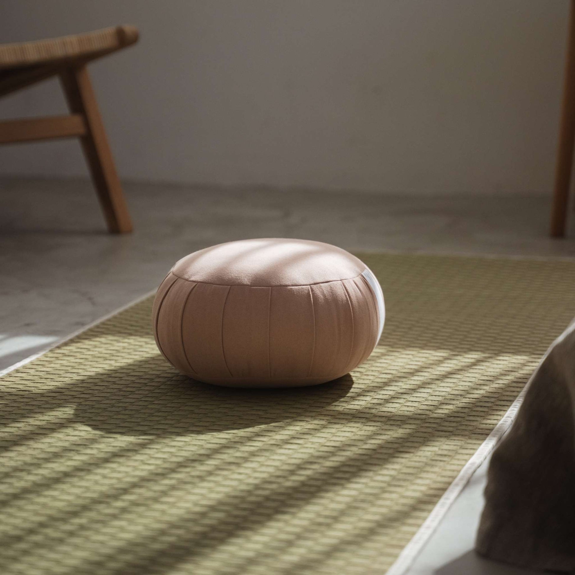 Coussin de méditation zafu rose fumé posé sur un tapis de tatami dans une pièce paisible avec des ombres de soleil.